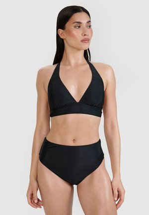 Kobieta w czarnym bikini z wiązaniem na szyi i wysokim stanem, stojąca na jednolitym jasnym tle, patrząca w prawo.