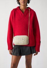 Maglione rosso a maglia con scollatura a V profonda e polsini a costine, abbinato a una borsa a tracolla dorata con dettagli del logo impresso e tracolla a catena.