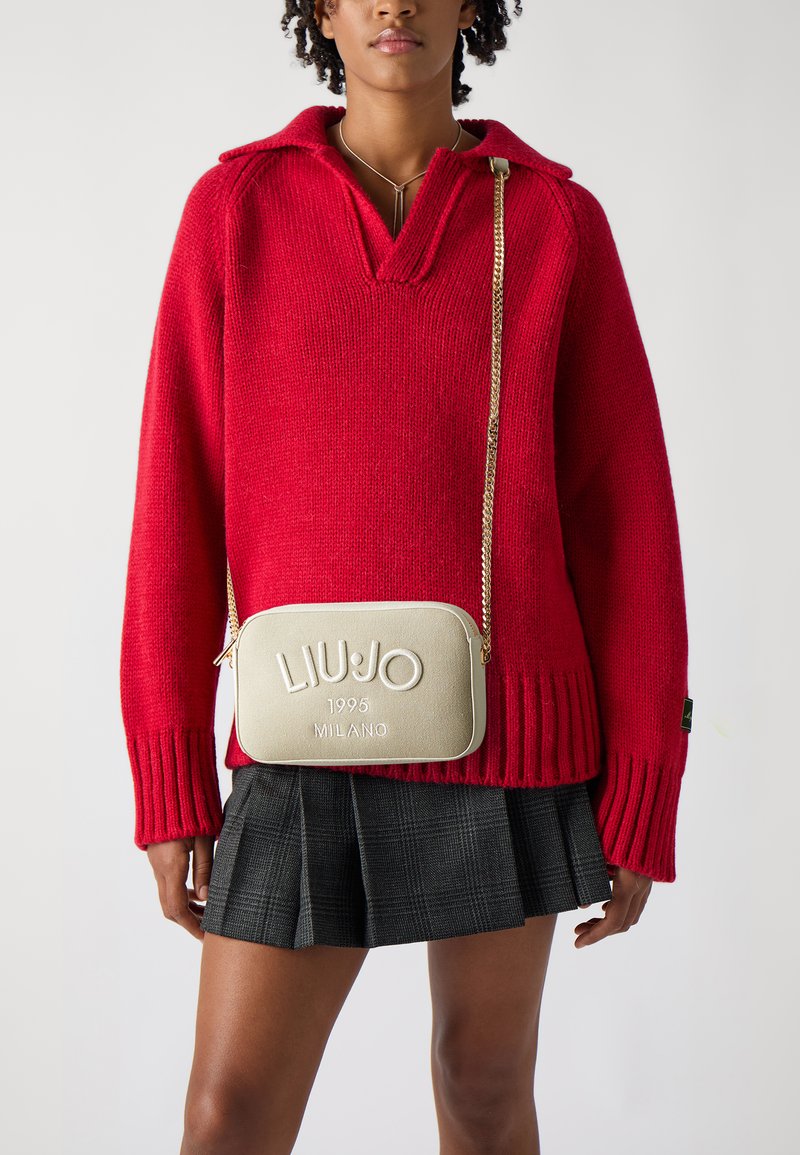 Maglione rosso a maglia con scollatura a V profonda e polsini a costine, abbinato a una borsa a tracolla dorata con dettagli del logo impresso e tracolla a catena.