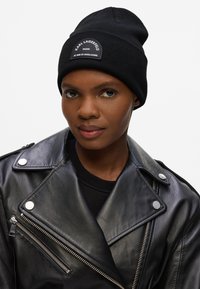 Gorro de punto negro con una superficie texturizada y un parche con logo. Se lleva con una chaqueta de cuero negra que cuenta con botones a presión metálicos y cremalleras.