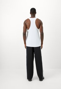 Mand iført en hvid ærmeløs tanktop og sorte løse bukser med hvide striber, stående overfor en ensfarvet hvid baggrund.