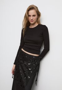 Črn dolg rokav s prilagojenim dizajnom, v kombinaciji s črno zavito krilo, ki ima okrasne kamenčke in teksturiran, večplastni videz.