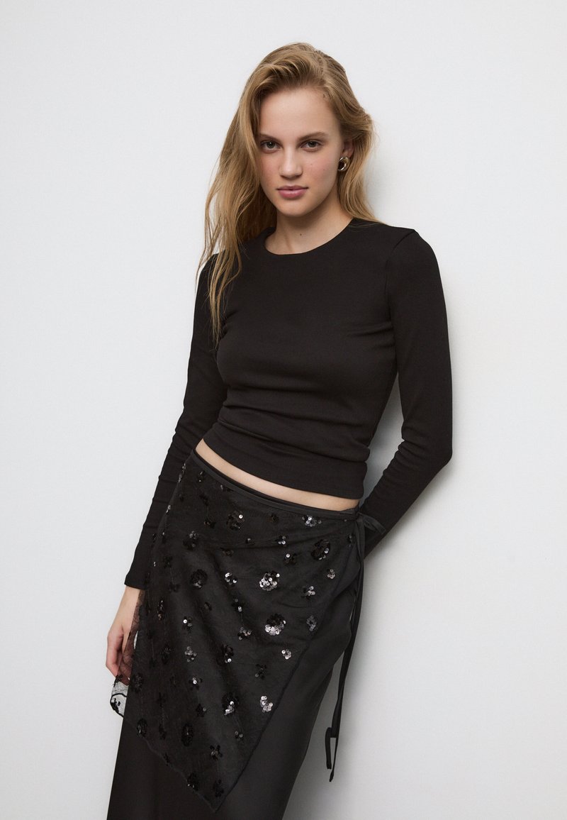 Črn dolg rokav s prilagojenim dizajnom, v kombinaciji s črno zavito krilo, ki ima okrasne kamenčke in teksturiran, večplastni videz.