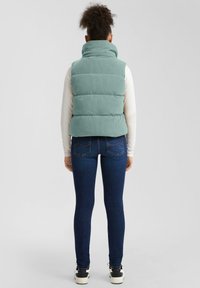 Žena stojaca chrbtom, oblečená v svetlozelenom zateplenom veste, bielej blúzke s dlhými rukávmi, tmavomodrých džínsoch a bielych teniskách.