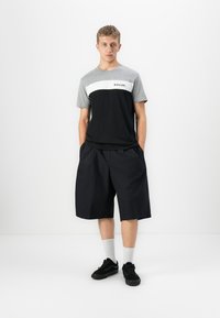 Ung mand står med hænderne i lommerne iført en grå og sort Rip Curl T-shirt, lange sorte shorts, hvide sokker og sorte sneakers.