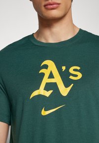 Zelené bavlnené tričko s veľkým žltým logom "A's" a Nike swoosh, s okrúhlym výstrihom a krátkymi rukávmi.