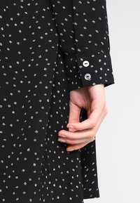 Tissu de chemise noir avec un motif à pois blancs ; présente des poignets boutonnés et une texture lisse ; main tenant le tissu près du poignet.