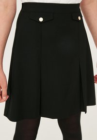 Jupe noire trapèze avec deux poches à rabat à l'avant, fermées par des boutons ronds dorés, portée avec des collants noirs et un haut blanc.