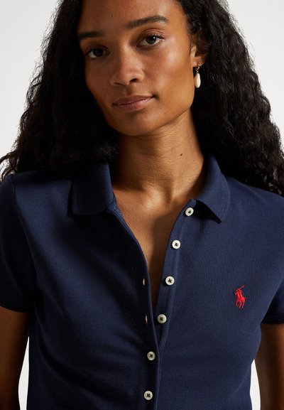 Marineblå polo med teksturert finish, fem hvite knapper og en rød brodert logo på venstre bryst. Korte ermer og krage.