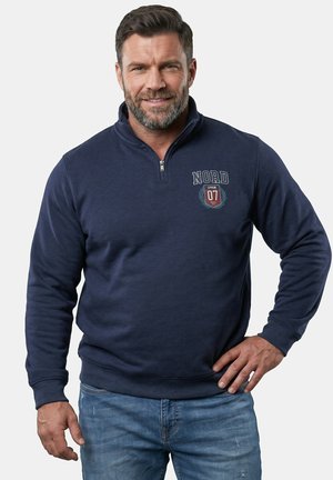 Skæggede mand iført en mørkeblå lynlåssweatshirt med "NORD 07" logo og afslappede blå jeans, smilende med den ene hånd på hoften.