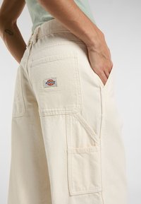Pantaloni cargo color crema con tessuto testurizzato, tasche laterali e posteriori, e una toppa con logo Dickies sulla tasca posteriore.