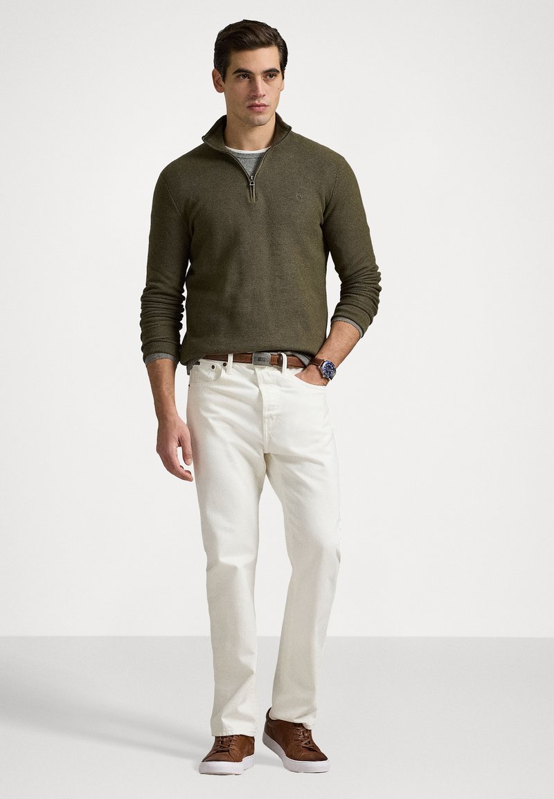 Maglione verde oliva con collo a zip, jeans bianchi e scarpe marroni. Presenta un tessuto lavorato e dettagli di cucitura sottili.