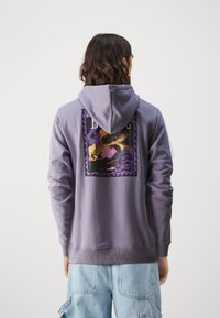 Fialová mikina s kapucňou s grafickým dizajnom v čiernej, oranžovej a fialovej na zadnej strane. Materiál je mäkký s rebrovanými manžetami a lemom.
