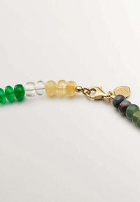 Collar de cuentas hecho de cuentas de vidrio verde, amarillo y transparente. Presenta un cierre de color dorado y una etiqueta redonda con un borde retorcido.