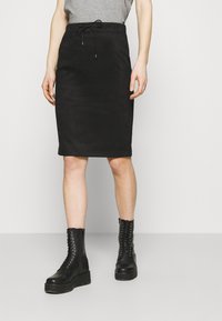 Jupe crayon en daim noir avec taille à cordon. Elle présente une coupe slim et une longueur jusqu'aux genoux, associée à des bottes montantes en dentelle noires.