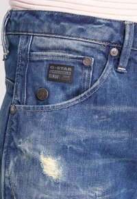 Jeans de mezclilla en azul claro con un acabado desgastado, que presentan una etiqueta negra, botones de metal y zonas desgastadas en el bolsillo delantero.