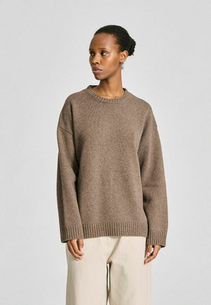 Femme debout, regardant vers la gauche, portant un pull en maille marron ample et un pantalon beige sur un fond gris clair uni.
