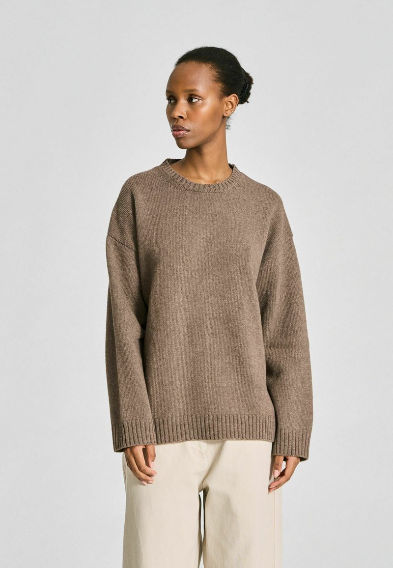 Femme debout, regardant vers la gauche, portant un pull en maille marron ample et un pantalon beige sur un fond gris clair uni.