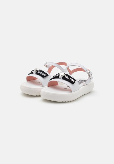 Sandalias blancas con tiras negras que presentan el logo 'Moschino', acentos en forma de corazón plateado y una suela texturizada. Forro interior de suave color rosa.