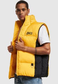 Pohodlná vesta s prednou časťou v žltej farbe a čierny chrbát. Má vysoký golier, zips a logo "SOUTH POLE" na hrudi. Odolný materiál.