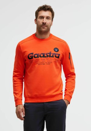 Man wearing bright orange Gaastra sweatshirt with navy print and patch on sleeve, paired with dark trousers, standing against white background.