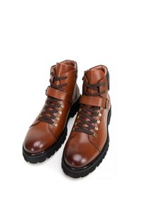 Botas de cuero marrón hasta el tobillo con cordones marrón oscuro, punteras redondeadas, suelas de goma y un detalle de hebilla en el lateral. Textura suave y diseño robusto.