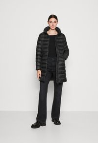 Zwarte pufferjas met rits aan de voorkant, capuchon en gestikte details, gedragen over een zwart top en gecombineerd met zwarte flared jeans en laarzen.