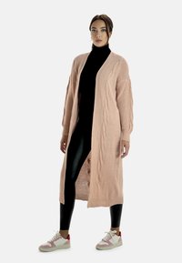 Lång, ljusrosa cardigan i ribbad stickning med kabeldetaljer längs framsidan. Kombinera med svarta leggings och vita sneakers med röda accenter.