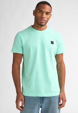Camiseta de algodón verde menta con cuello redondo, mangas cortas y una pequeña etiqueta negra en el pecho izquierdo. Se lleva con unos vaqueros azul claro.