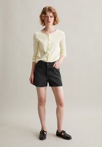 Lichtgele geribbelde cardigan met knopen, gecombineerd met zwarte denim shorts. Het model draagt zwarte instappers en staat tegen een neutrale achtergrond.