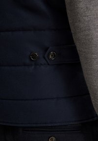 Granatowa kamizelka o gładkiej fakturze, z dwoma zapięciami na guziki i dopasowanym krojem, noszona pod jasnoszarym materiałem.