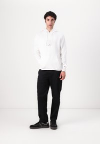Hvit hettegenser med kengurulomme og trykt logo, kombinert med svarte bukser og svarte joggesko med hvite detaljer.