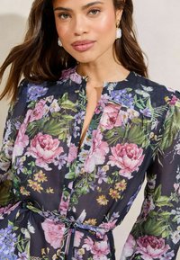 Vloeiende, doorschijnende bloemenjurk met roze en paarse bloesems op een zwarte achtergrond, voorzien van een mandarin kraag en een ceintuur in de taille.