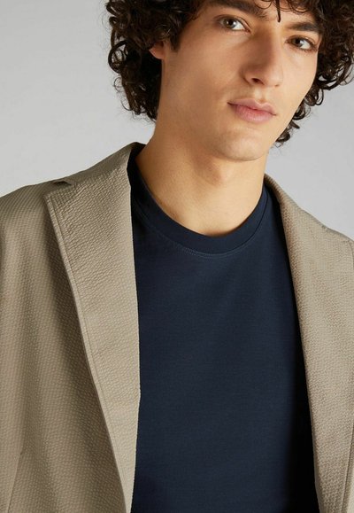 Blazer beige texturé sur un t-shirt à col rond bleu marine foncé, doté d'un col cranté et de détails de couture subtils.