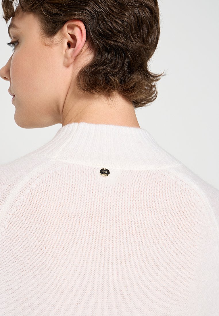 Suéter de punto blanco con un acabado texturizado, que presenta un cuello acanalado y un pequeño botón negro como detalle en la parte posterior del escote.