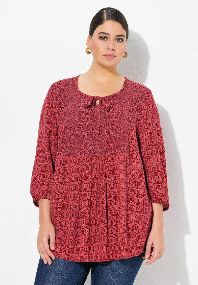 Rote Bluse mit strukturiertem Muster, runder Halslinie und Schlitzdetail. Sie hat 3/4-lange Ärmel und ein lockeres, gerafftes Design.