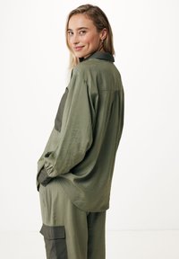 Maglietta a maniche lunghe verde oliva con colletto e tasche laterali. Presenta una tasca a toppa di colore scuro in contrasto realizzata in un tessuto morbido e testurizzato.