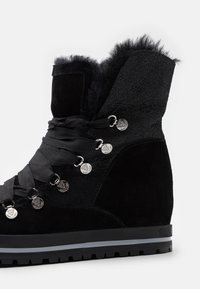 Bottine noire à la cheville avec doublure en fourrure, lacets larges en ruban, œillets métalliques avec logo, dessus texturé, et semelle en caoutchouc avec une bande grise.