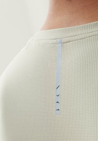 Gaiši bežs teksturēts T-krekls ar sietveida rakstu, ar zilu birku "BRN" tuvu apkakles aizmugurē.