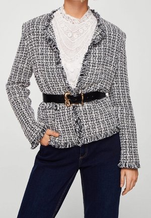 Femme portant un blazer à franges en tweed noir et blanc avec une ceinture noire, un chemisier blanc en dentelle et un jean bleu foncé, debout avec une main dans la poche.