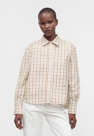 Personne portant une chemise à carreaux beige et marron à boutons avec manches longues et un pantalon blanc sur un fond uni lumineux.