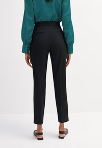 Czarne dopasowane spodnie o gładkiej fakturze, z prostymi nogawkami, frontowymi zakładkami i wysokim stanem. Dyskretne tylne kieszenie widoczne.