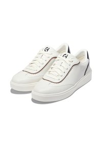 Ett par vita lågskor med krämfärgade snörning, bruna sömmar runt ögla, och svarta detaljer på hälen, visade på vit bakgrund.