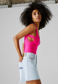 Γυναίκα με μαλλιά σγουρά, που φοράει φωτεινό ροζ αμάνικο τοπ και φούστα από ανοιχτό μπλε τζιν, στέκεται με τα χέρια στις πίσω τσέπες.