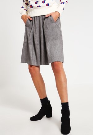 Jupe en suède gris avec des plis et des poches latérales ; présente une section inférieure texturée avec des détails perforés. Longueur cheville, associée à des bottines noires.