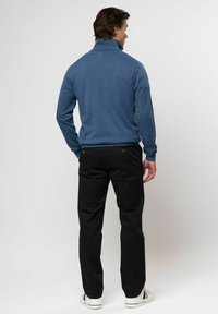 Vyras žiūrintis į kitą pusę, vilkintis mėlynį golfą, juodas kelnes ir baltus sportinius batelius su raudonomis ir mėlynomis juostelėmis, priešais vienspalvį foną.