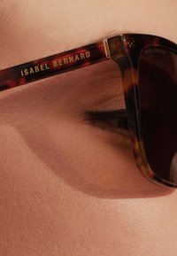Des lunettes de soleil en écaille de tortue présentent une monture épaisse avec la marque dorée "ISABEL BERNARD". Des verres sombres partiellement obstrués par un cil.