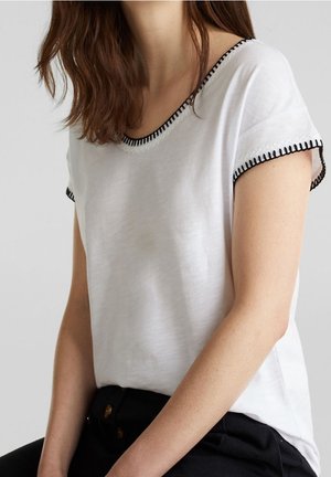 Kobieta w białym topie z krótkim rękawem z czarnym przeszyciem na dekolcie i krawędziach rękawów, w połączeniu z czarnymi spodniami.