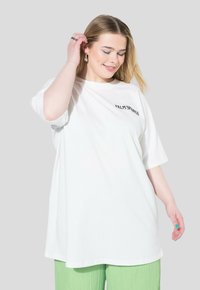 Camiseta blanca de gran tamaño en suave tejido de algodón, con texto negro "PALM SPRINGS" en la parte delantera, combinada con pantalones plisados de color verde claro.