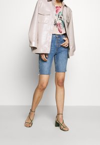 Veste chemise en simili cuir rose clair, t-shirt avec motif floral, shorts en denim Bermuda et sandales à talons à lanières vertes.